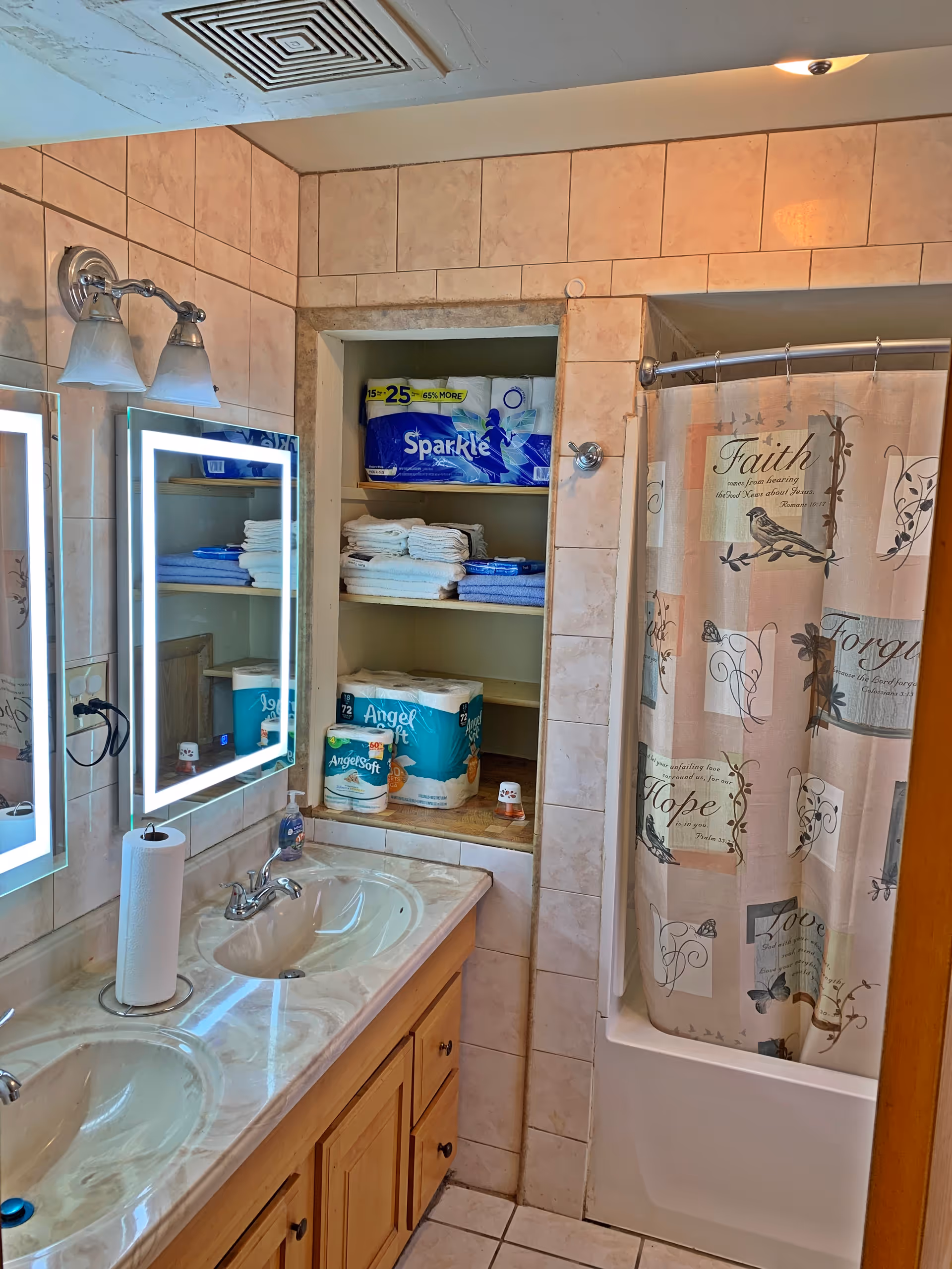 A bathroom with a double sink vanity featuring a marble countertop and illuminated mirrors above each sink. There is a built-in shelving unit stocked with towels and toilet paper. To the right, there is a bathtub with a shower curtain decorated with words like Faith, Hope, and Love along with bird and floral designs. The walls and floor are tiled in a beige color.