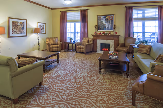 Spacious furnished communal living room with sofas, armchairs, coffee tables, a fireplace, and windows with curtains.