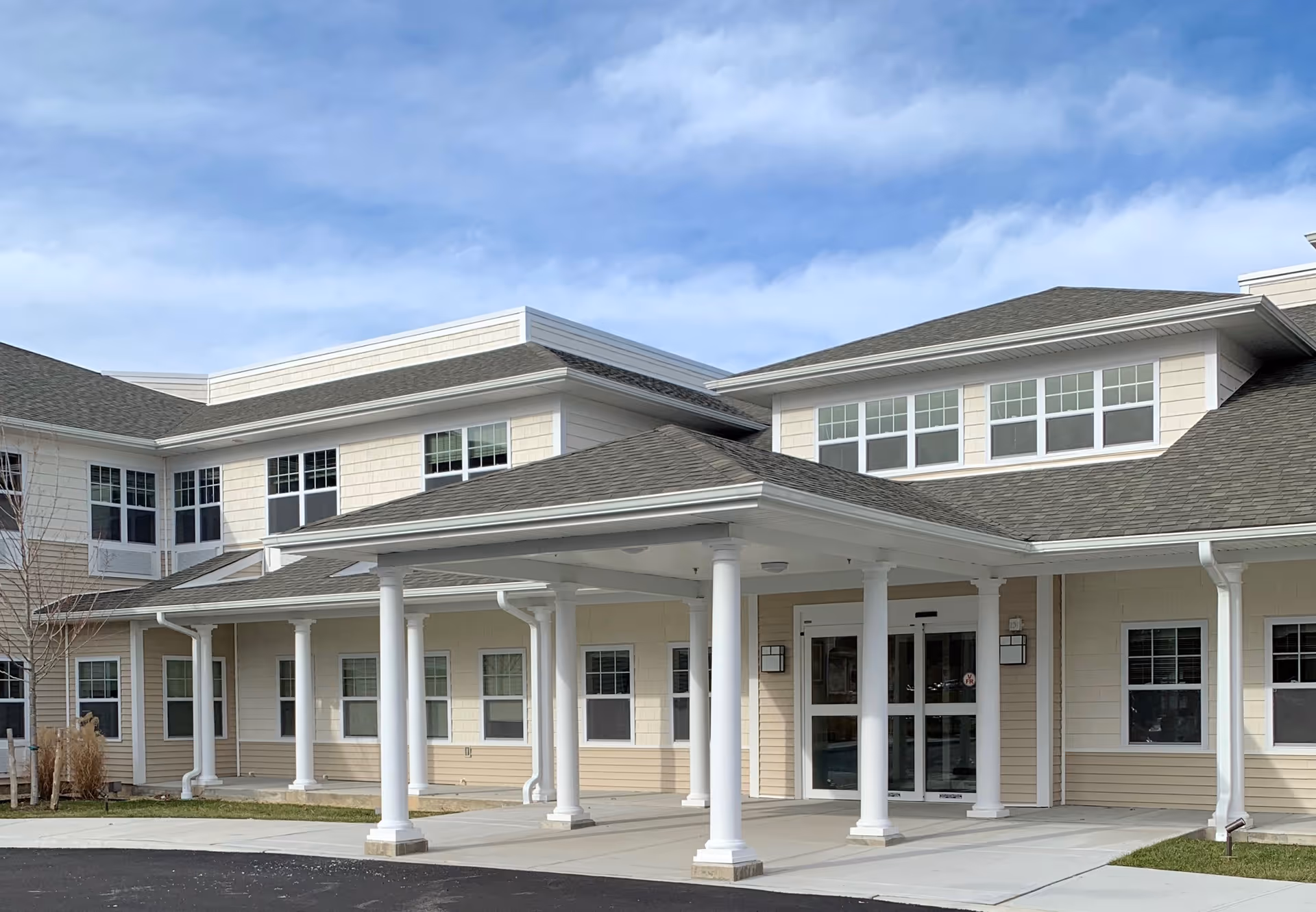 Exterior view of a two-story senior living facility building with beige siding and white trim. The entrance features a covered porch supported by white columns, with glass double doors. The sky is partly cloudy.