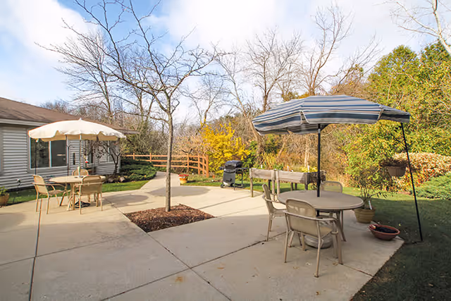 Concrete patio with round tables, chairs and umbrellas adjacent to a single-story building and landscaped yard.