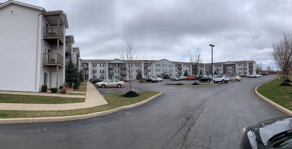 Wide exterior view of a three-story senior apartment complex with parked cars and an overcast sky.