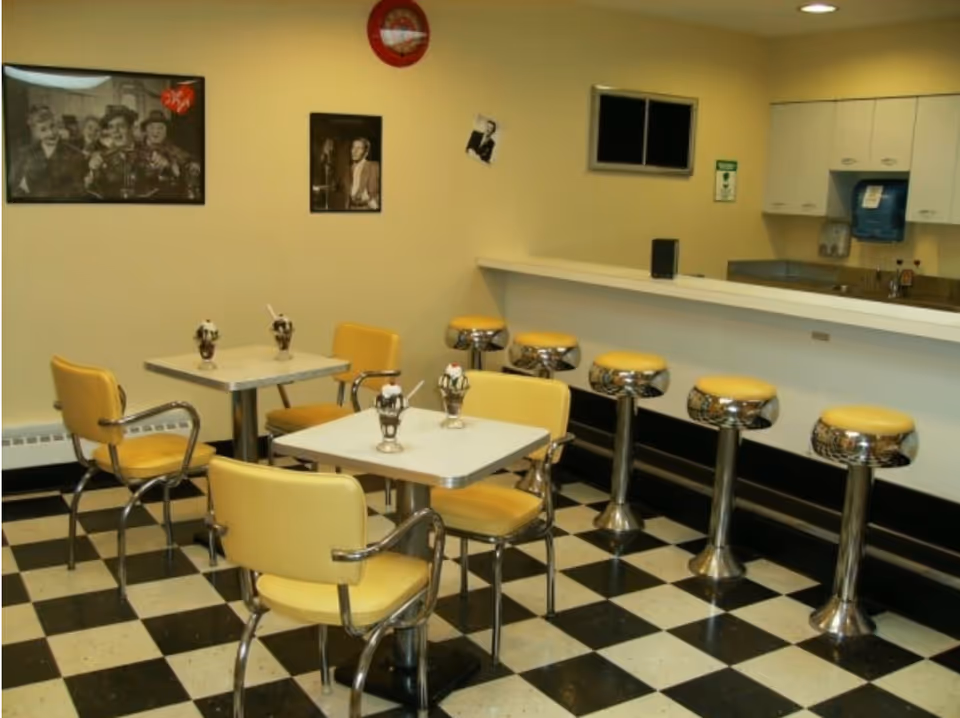 Retro diner-style dining area with yellow chairs, chrome counter stools, checkered floor and small tables with dessert dishes.