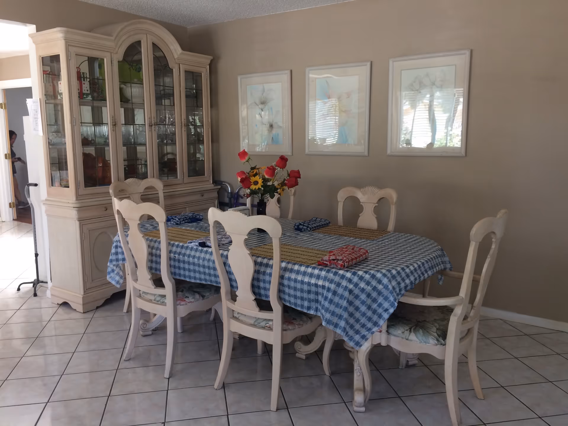 A dining room with a blue-checkered tablecloth on a table surrounded by six white chairs, a china cabinet, framed artwork on the wall, and a vase of flowers.