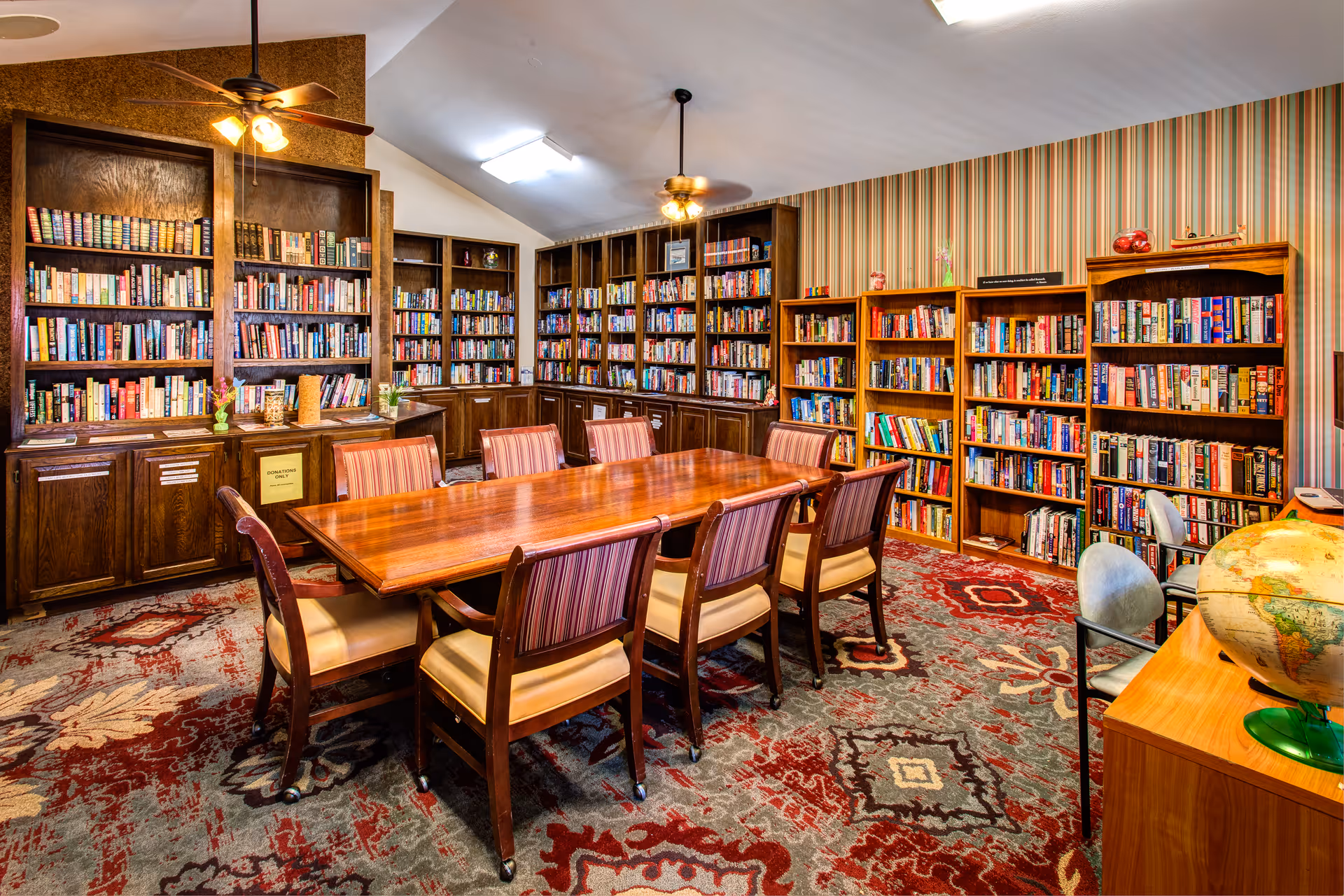 A cozy library room with wooden bookshelves filled with books lining the walls. In the center, there is a large wooden table surrounded by eight chairs with striped upholstery. The room has a patterned carpet with red, gray, and beige tones, two ceiling fans with lights, and a globe on a wooden desk in the foreground.
