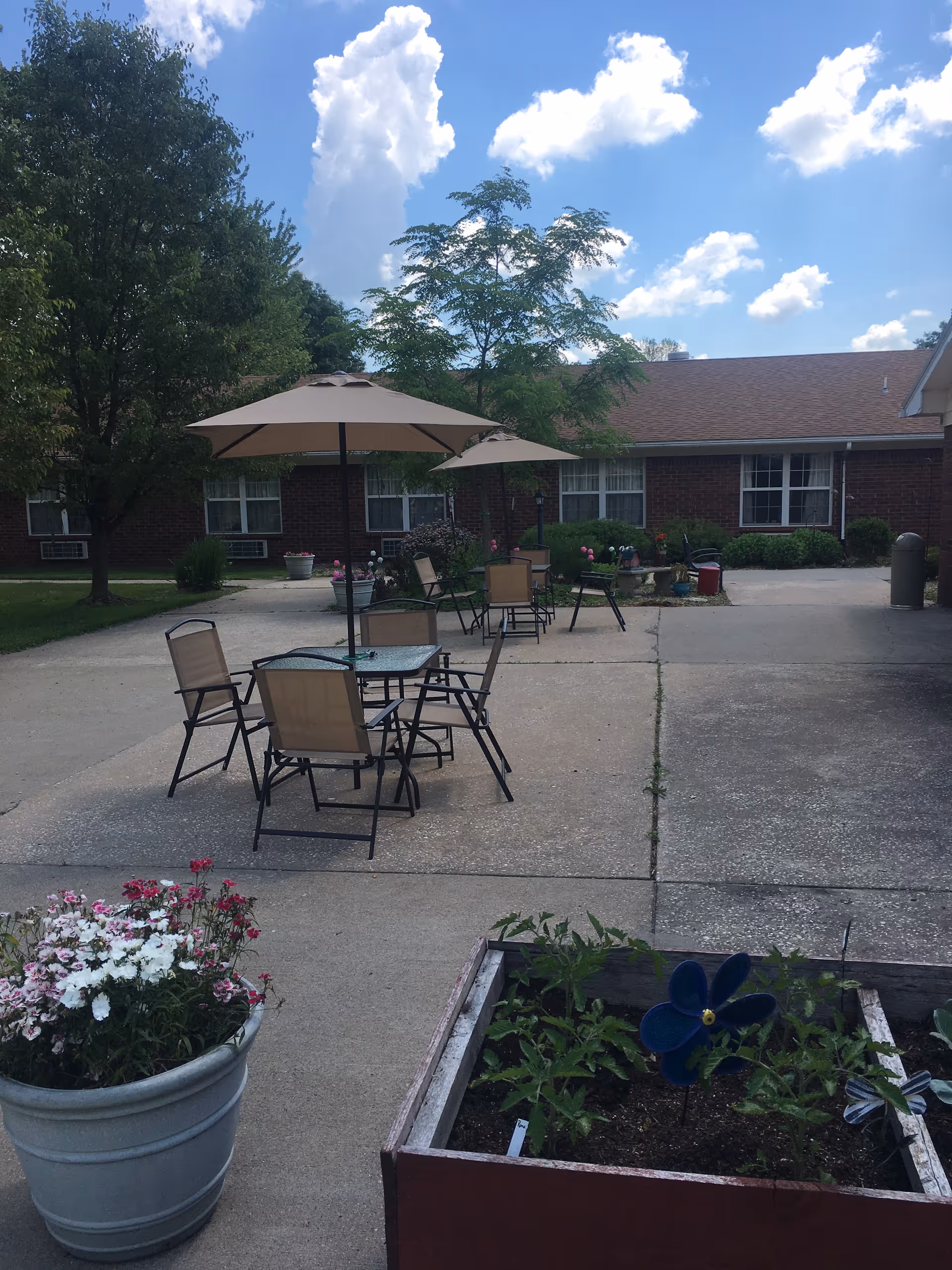 Outdoor courtyard with patio tables and umbrellas, potted flowers, and a one-story brick building in the background.