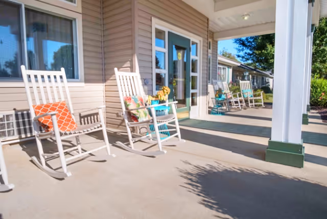A covered outdoor patio area at Emerald Place featuring several white rocking chairs with colorful cushions. The patio is adjacent to a beige building with large windows and a glass door. There are green mats on the concrete floor and some potted plants near the chairs. Trees and shrubs are visible in the background.