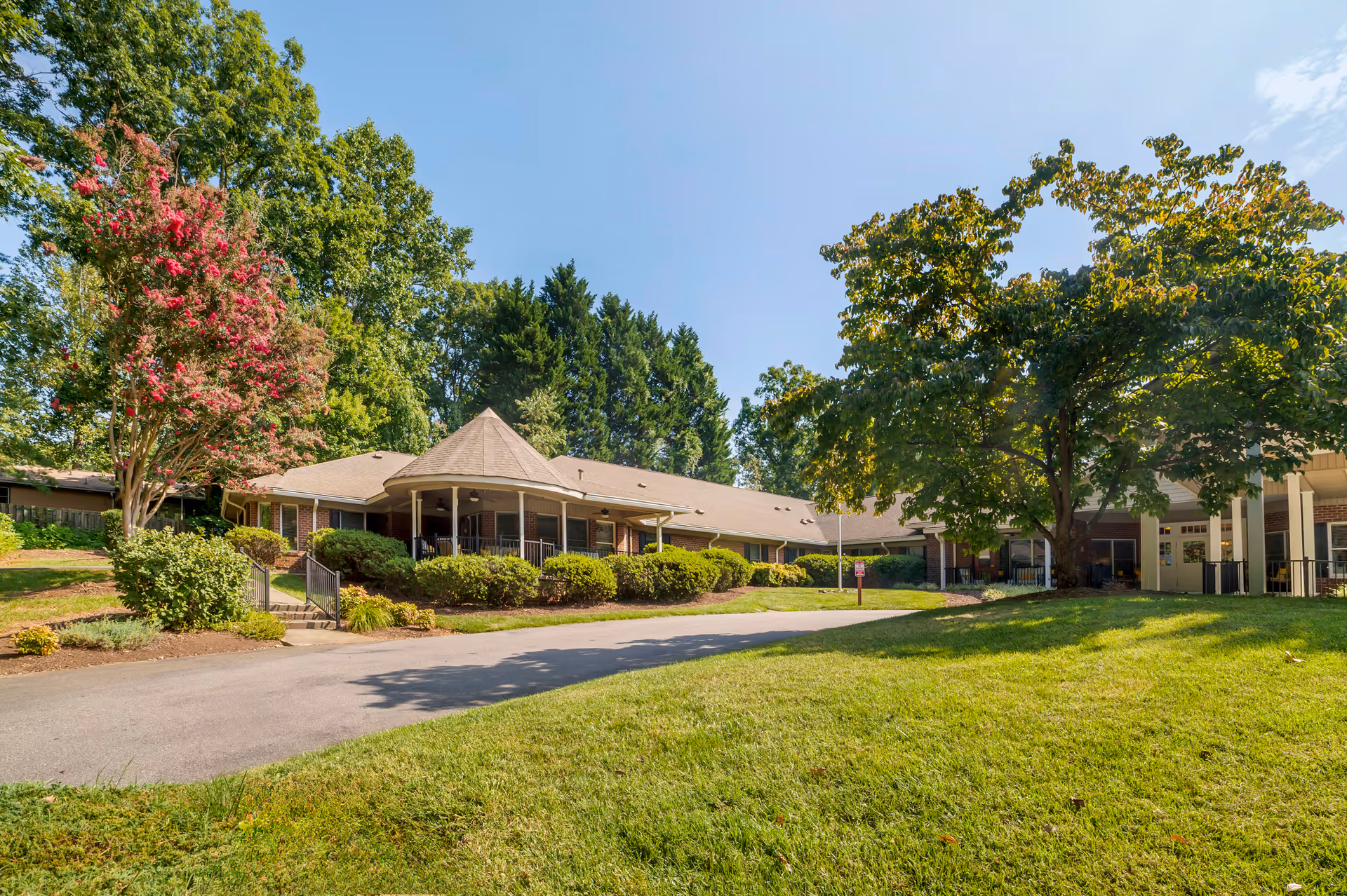 Exterior view of Brookdale Asheville Overlook senior living facility showing a single-story building with a covered porch, surrounded by green trees, bushes, and a well-maintained lawn under a clear blue sky.