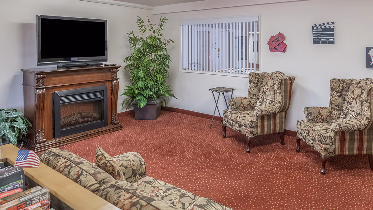 A cozy senior living community lounge area with a flat-screen TV mounted on a wooden fireplace mantel, two patterned armchairs, a matching sofa, a small side table, and potted plants. The room has a red carpet and white walls with decorative wall art including movie tickets and a clapperboard.