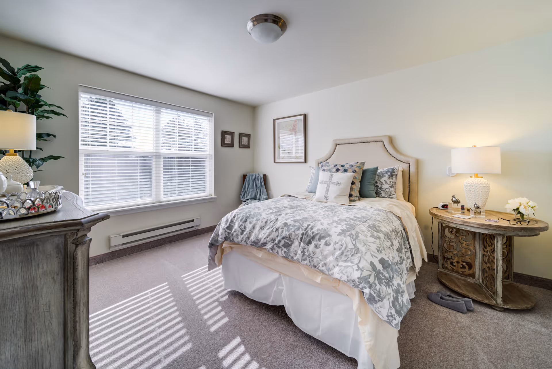 A bright and cozy bedroom with a large window letting in natural light. The room features a bed with a beige upholstered headboard, floral-patterned bedding, and multiple decorative pillows. There is a wooden nightstand with a white lamp, flowers, and personal items, as well as a wooden dresser with a lamp and decorative tray. The walls are light-colored, and the carpet is a soft neutral tone.