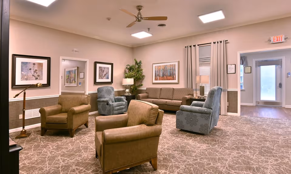 A cozy living room area with a mix of brown and blue upholstered armchairs and a brown sofa. The room has beige walls with framed artwork, a ceiling fan, a floor lamp, a table lamp, and a potted plant. There is a patterned carpet on the floor and an exit door with a window covered by curtains.