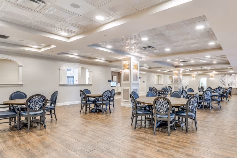 A spacious dining room with multiple round wooden tables and elegant chairs featuring patterned upholstery. The room has a light-colored ceiling with recessed lighting and decorative ceiling panels. The floor is covered with wood-like tiles, and the walls are painted in a soft neutral tone with arched openings and decorative columns.