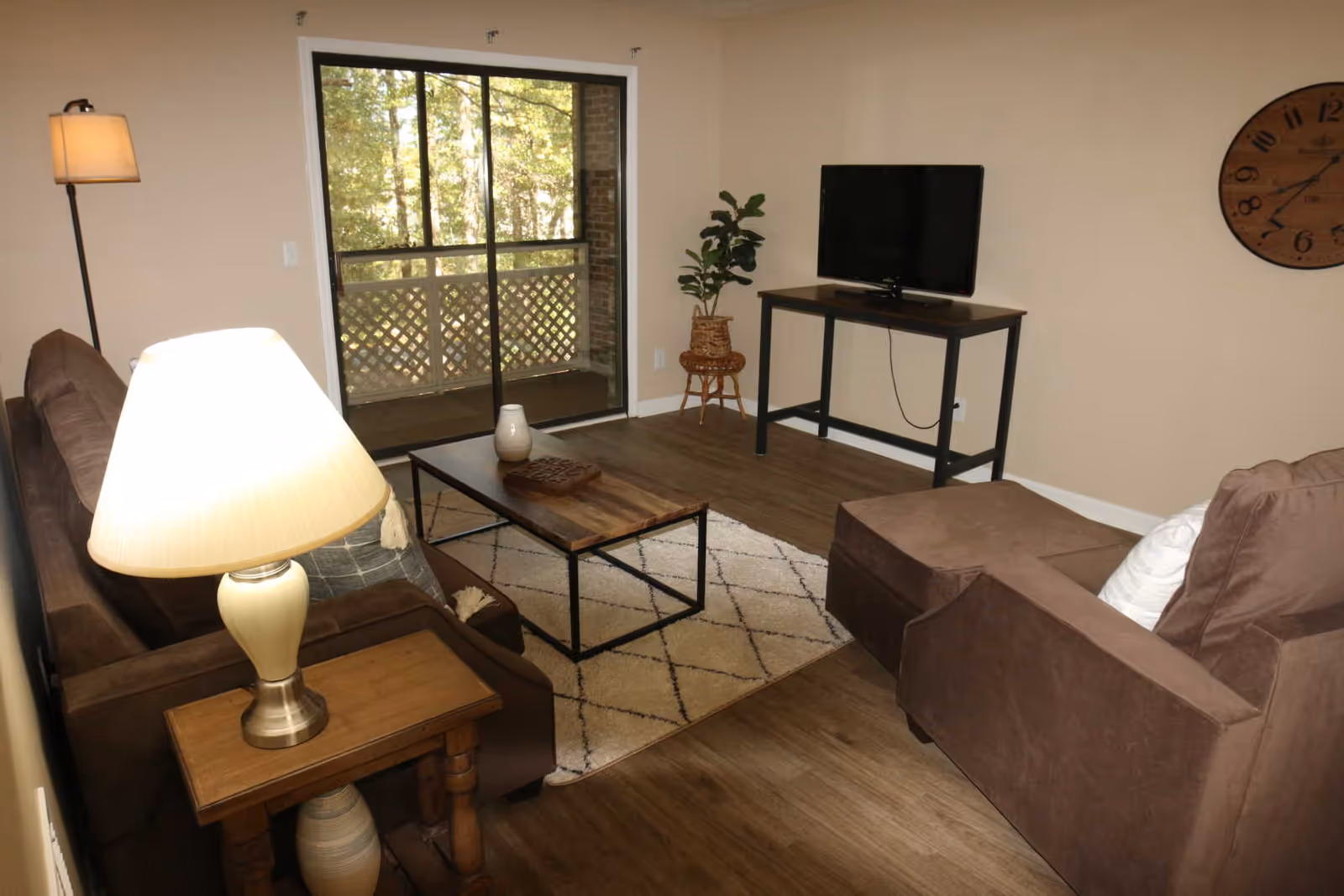 A cozy living room with brown sofas, a wooden coffee table on a patterned rug, a floor lamp, a table lamp on a side table, a flat-screen TV on a black metal stand, a large wall clock, a potted plant, and a sliding glass door leading to a balcony with a lattice railing and trees visible outside.