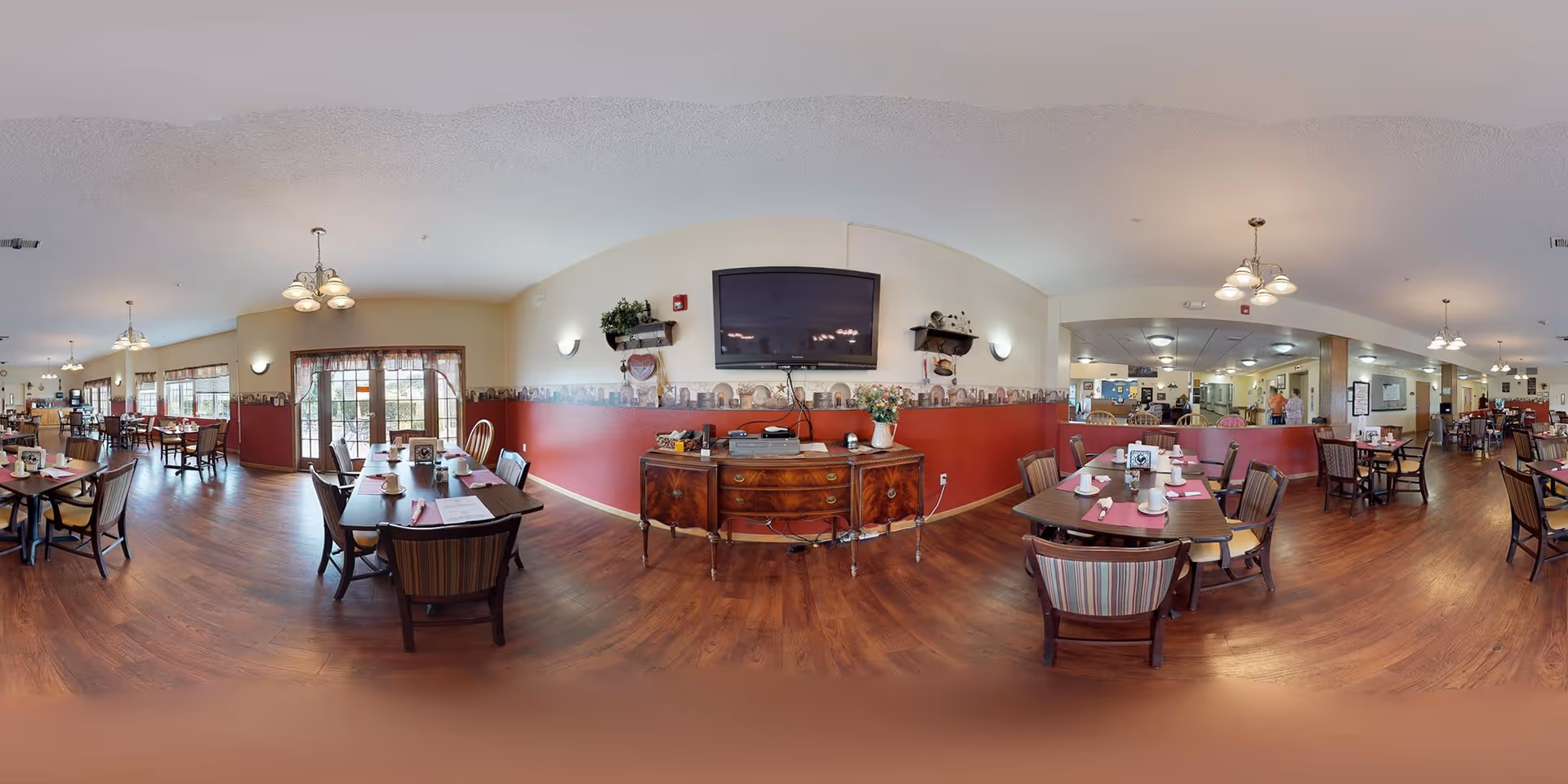 A spacious dining room in a senior living facility with multiple wooden tables and chairs arranged neatly. The tables are set with placemats, napkins, and cups. The room features warm wood flooring, cream and red walls with decorative border trim, and several ceiling light fixtures. A large flat-screen TV is mounted on the wall above a wooden sideboard with decorative items and plants. Large windows and glass doors allow natural light to enter the room.