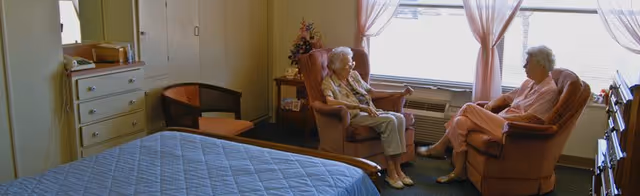 Two elderly women sitting and talking in a cozy room with a bed, dresser, and large window with pink curtains letting in natural light.
