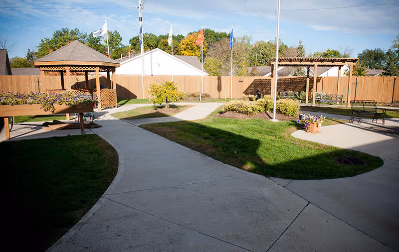 Outdoor courtyard area with paved walkways, green grass, flower beds, wooden gazebos, benches, and several flagpoles with flags against a blue sky and trees in the background.