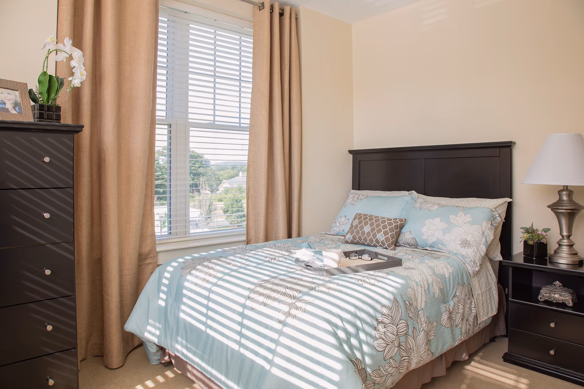 Sunlit bedroom with a floral-covered bed, dark wood headboard, matching dresser and nightstand beside a window with blinds and beige curtains.