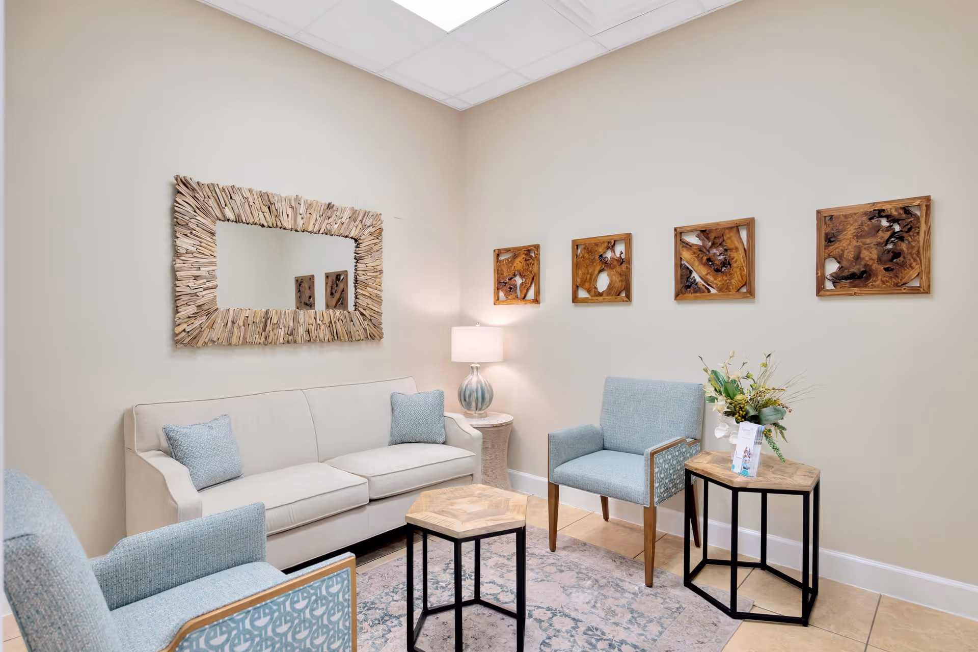 A cozy sitting area with a beige sofa and two light blue armchairs arranged around two small wooden tables. A large rectangular mirror with a driftwood frame hangs on the wall above the sofa. Four square wooden art pieces are displayed on the adjacent wall. A table lamp with a glass base sits on a small side table next to the sofa. The room has beige walls and tiled flooring with a patterned area rug.