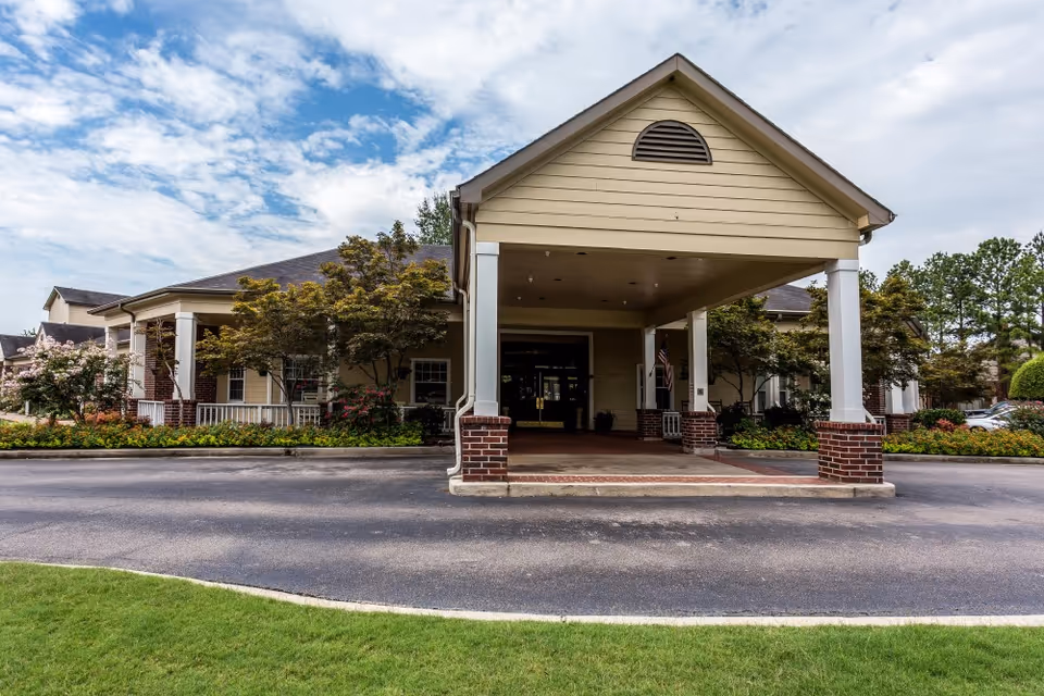 The front entrance of Hearthstone Assisted Living, featuring a covered porch with columns, landscaped gardens, and a paved driveway.