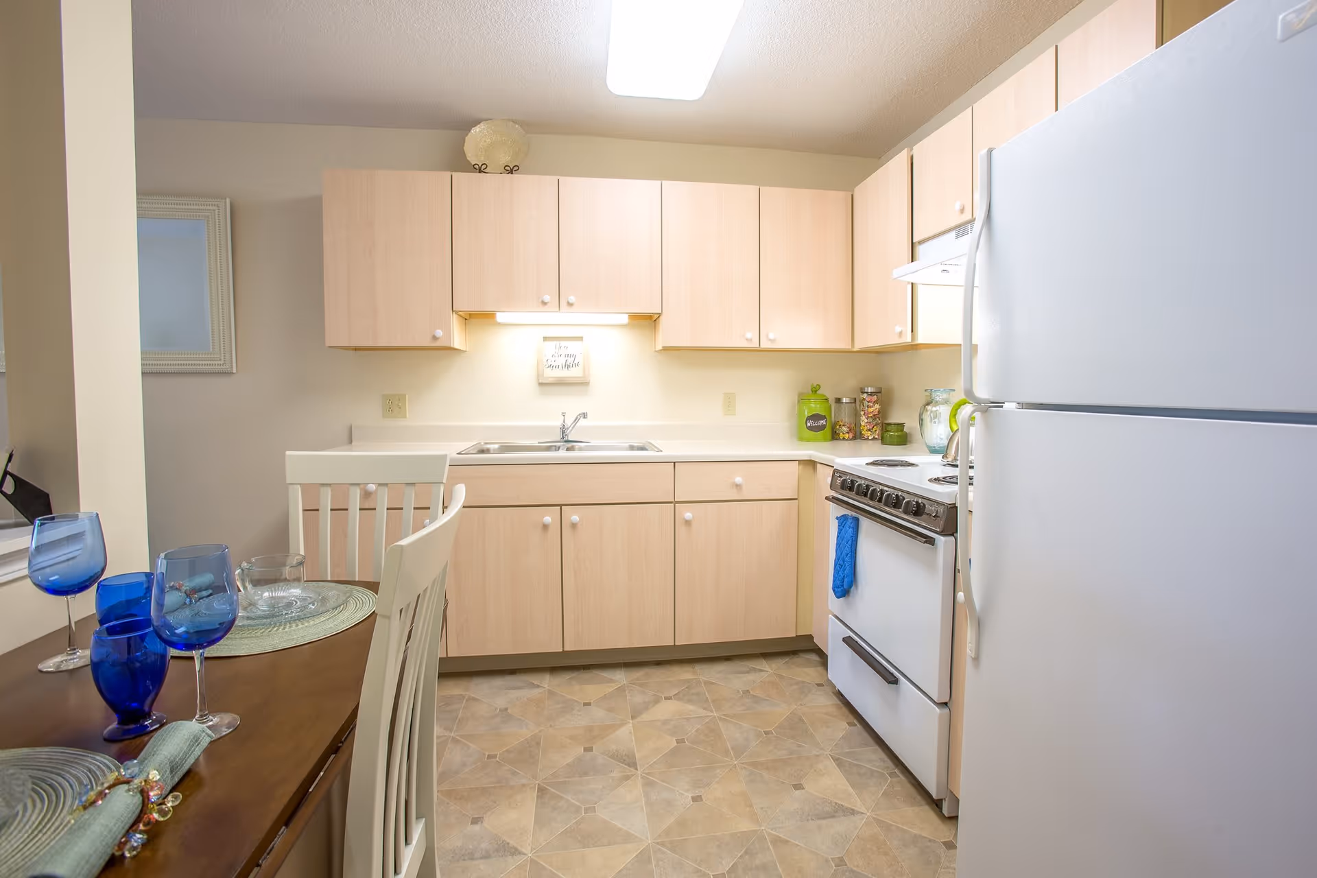 A compact kitchen with light wood cabinets, a white refrigerator, a white stove with a blue towel hanging on the handle, and a double sink. There is a small dining table with white chairs, set with blue glasses and placemats. The floor has a patterned tile design, and the kitchen is well-lit with a ceiling light and under-cabinet lighting.