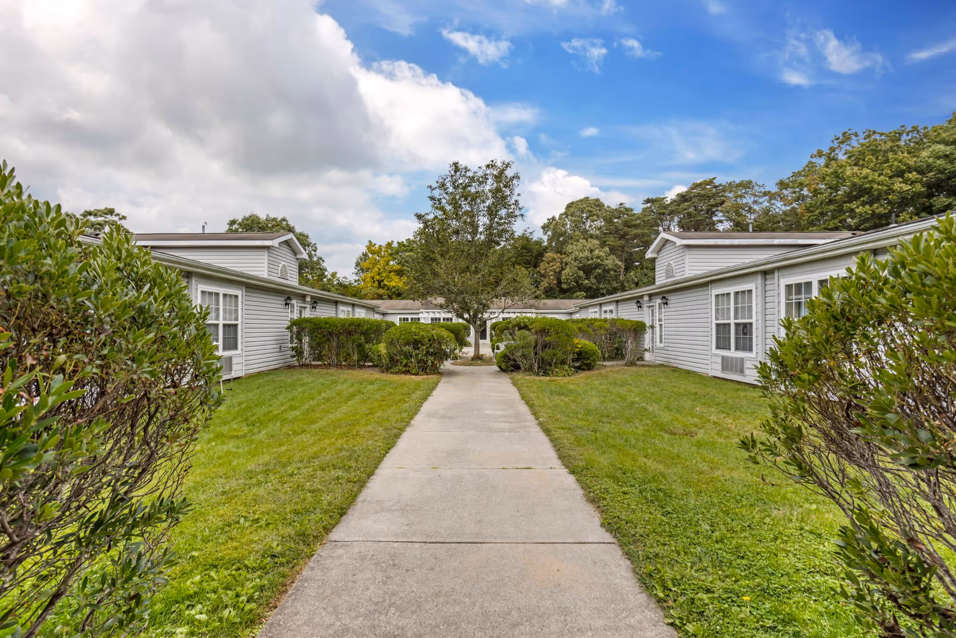 A concrete walkway flanked by green grass and trimmed bushes leads to a courtyard area with a tree in the center, surrounded by single-story gray buildings with white trim under a partly cloudy sky.