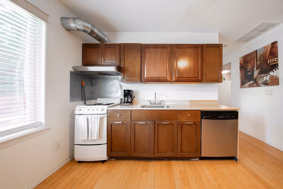 A kitchen with wooden cabinets, a white stove with a towel hanging on the handle, a coffee maker on the countertop, a stainless steel dishwasher, and a window with blinds letting in natural light. There is a painting on the wall to the right and light wood flooring.