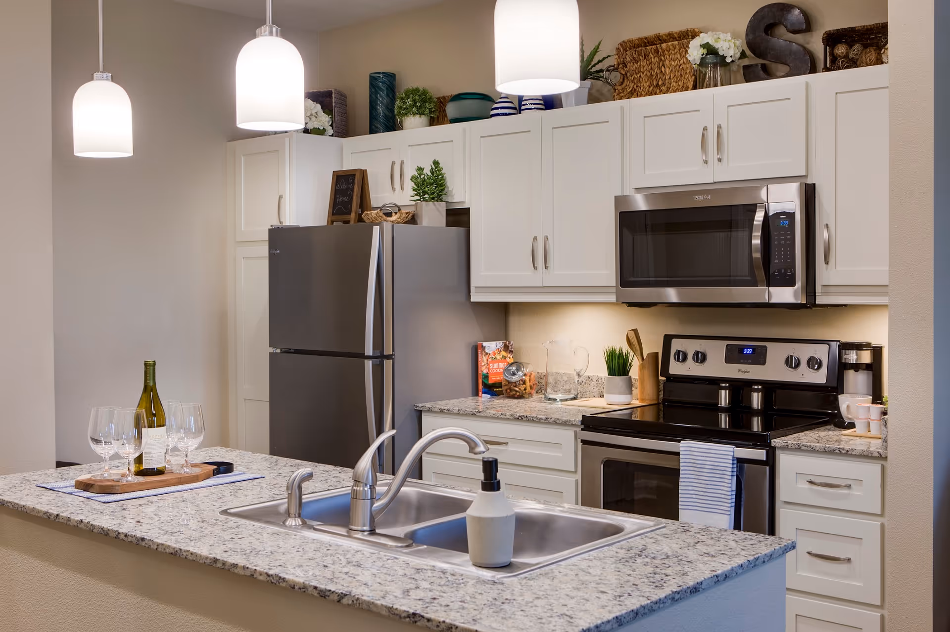 Modern kitchen with white cabinets, granite countertops, stainless steel refrigerator, oven, and microwave. A double sink with a soap dispenser is installed in the kitchen island, which also has a tray with a bottle of wine and three wine glasses. Pendant lights hang above the island, and decorative items are placed on top of the cabinets.