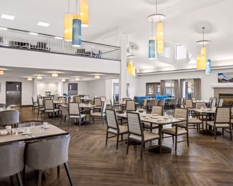Spacious dining room in a senior living facility with multiple tables and chairs arranged neatly. The room features high ceilings with modern hanging light fixtures in yellow and blue tones. There is a fireplace on the right side and large windows with curtains allowing natural light to enter. The floor has a wood pattern, and there is a mezzanine level with additional seating visible above.