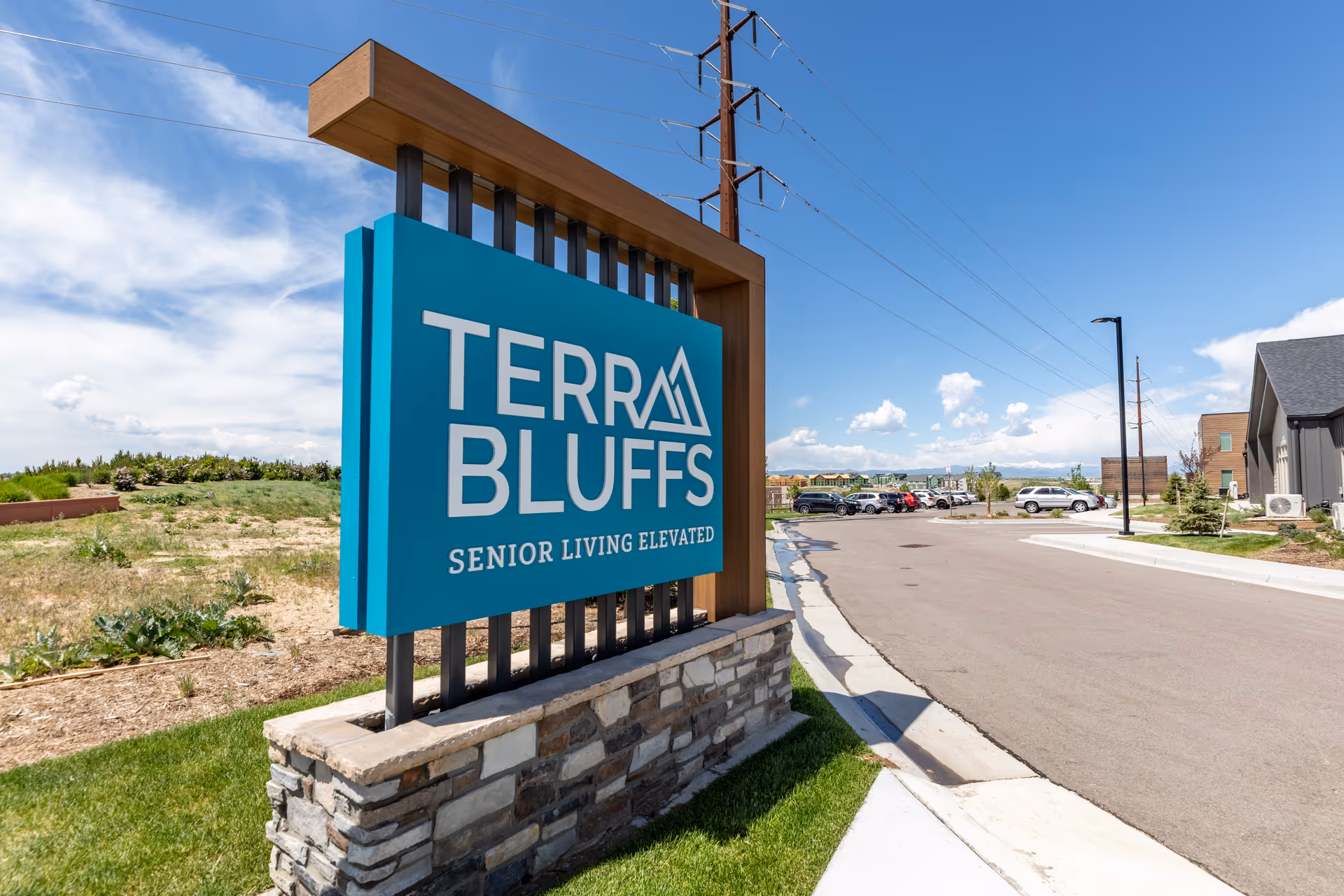 Outdoor view of a large blue sign with the text 'Terra Bluffs Senior Living Elevated' mounted on a stone and wood base, with a parking lot, buildings, and a partly cloudy sky in the background.