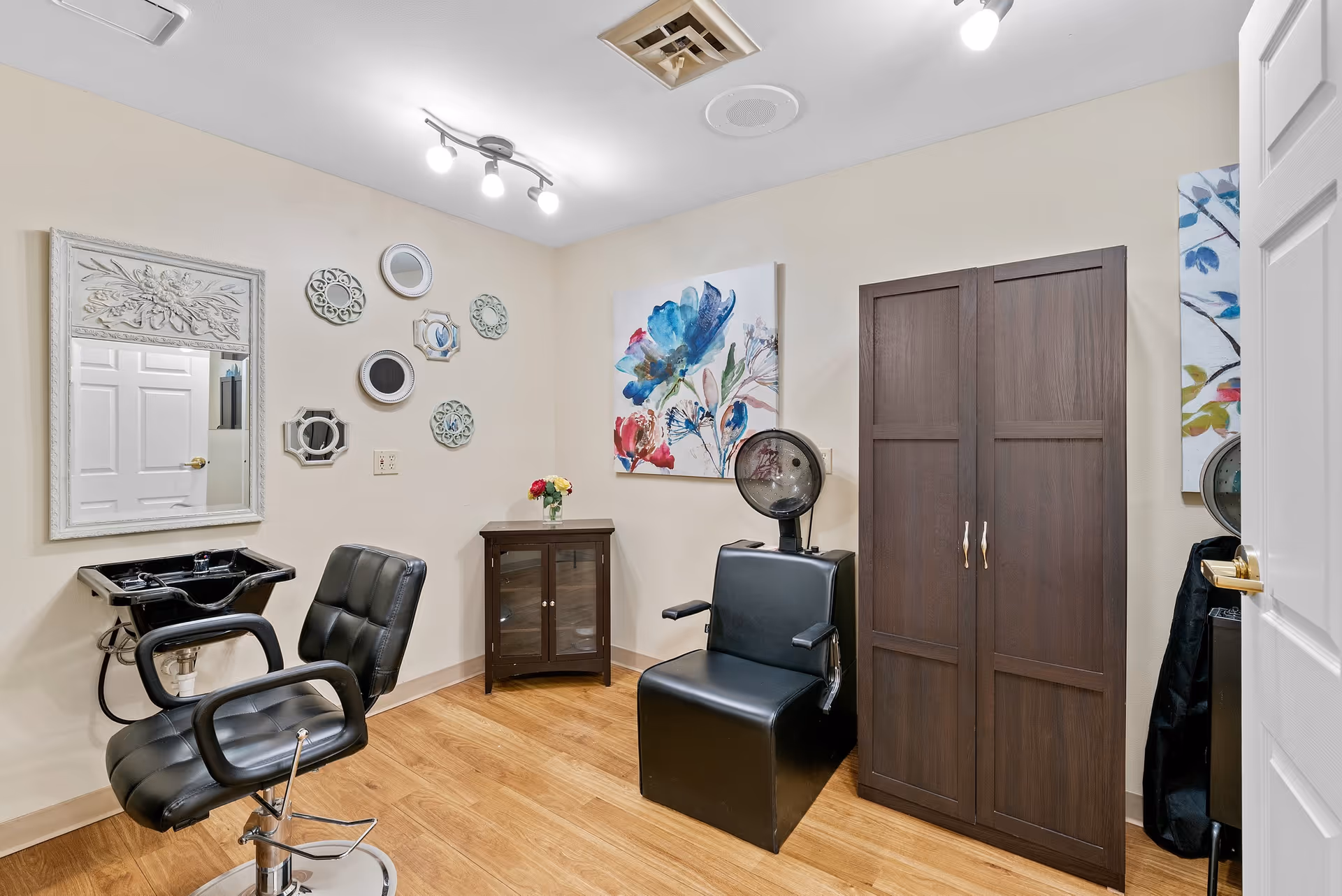 A small salon room with two black salon chairs, one in front of a black sink and mirror, and the other under a hair dryer. The room has light beige walls decorated with a floral painting and several small decorative mirrors. There is a dark wooden cabinet and a small table with a vase of flowers. The floor is wood, and the ceiling has a light fixture with three bulbs.
