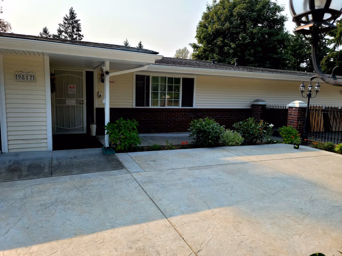 Exterior view of a single-story building with white siding and a brick lower half. There is a covered entrance with a white door and a house number plaque displaying 9817. The area in front of the building is paved with concrete and there are some green shrubs and plants along the building's foundation. A black metal fence and lamp posts are visible on the right side, with trees in the background.