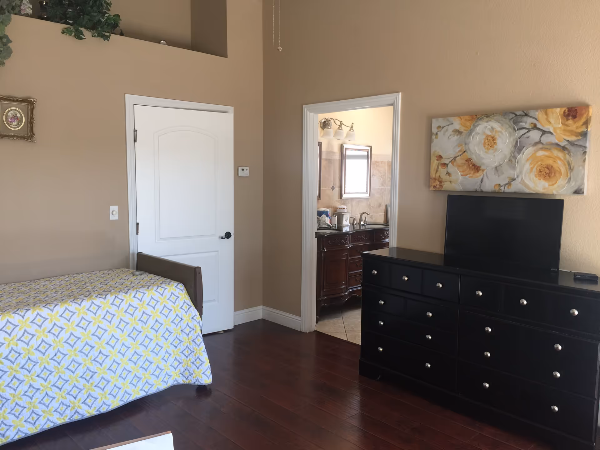 A bedroom with a single bed covered with a patterned yellow and gray bedspread, a black dresser with a flat-screen TV on top, and a floral painting hanging above the dresser. There is a door leading to a bathroom with a vanity and mirror visible. The walls are beige and the floor is dark wood.
