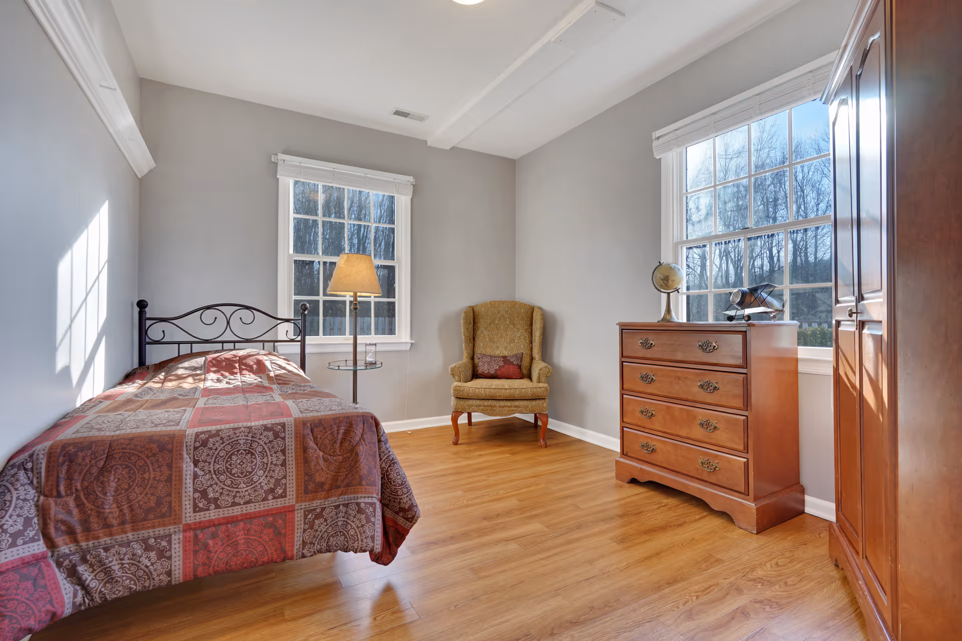 A bright and cozy bedroom with light gray walls and wooden flooring. The room features a single bed with a patterned quilt, a tall floor lamp beside the bed, an upholstered armchair with a decorative pillow, a wooden dresser with a globe and a model airplane on top, and a wooden wardrobe. Two large windows allow natural light to fill the room.