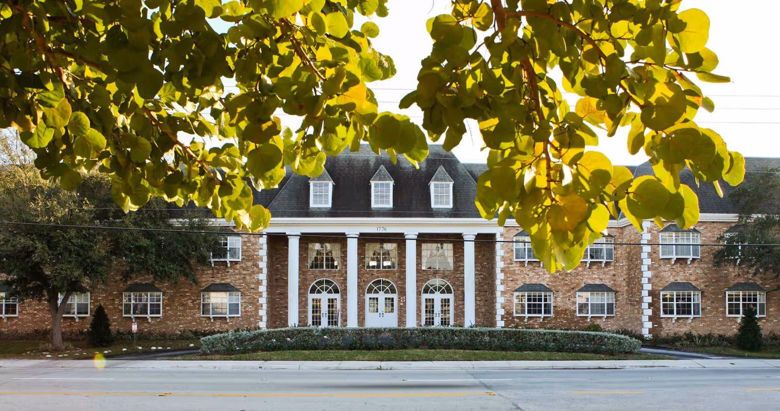Front exterior view of a large brick building with white columns and multiple windows, partially framed by green leafy tree branches.