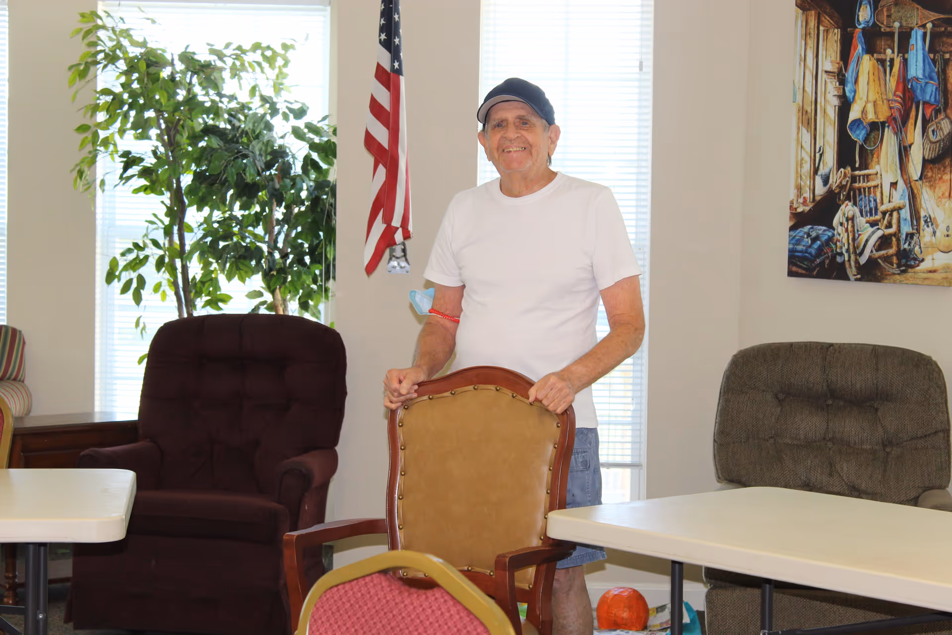 An elderly man wearing a white t-shirt, blue shorts, and a black cap stands indoors holding the back of a brown chair. Behind him are two large windows with blinds, an American flag, a large green plant, and two cushioned armchairs. A colorful painting hangs on the wall to the right.