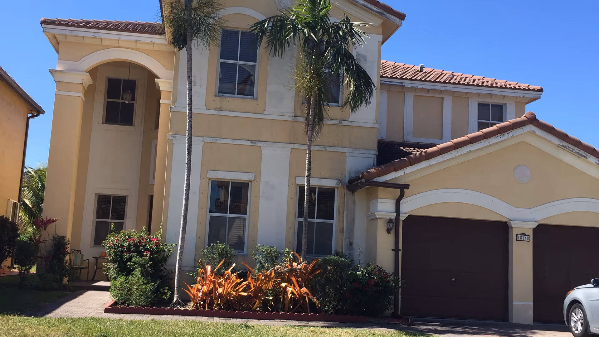 Two-story yellow stucco house with a tiled roof, two-car garage, palm trees and front landscaping.