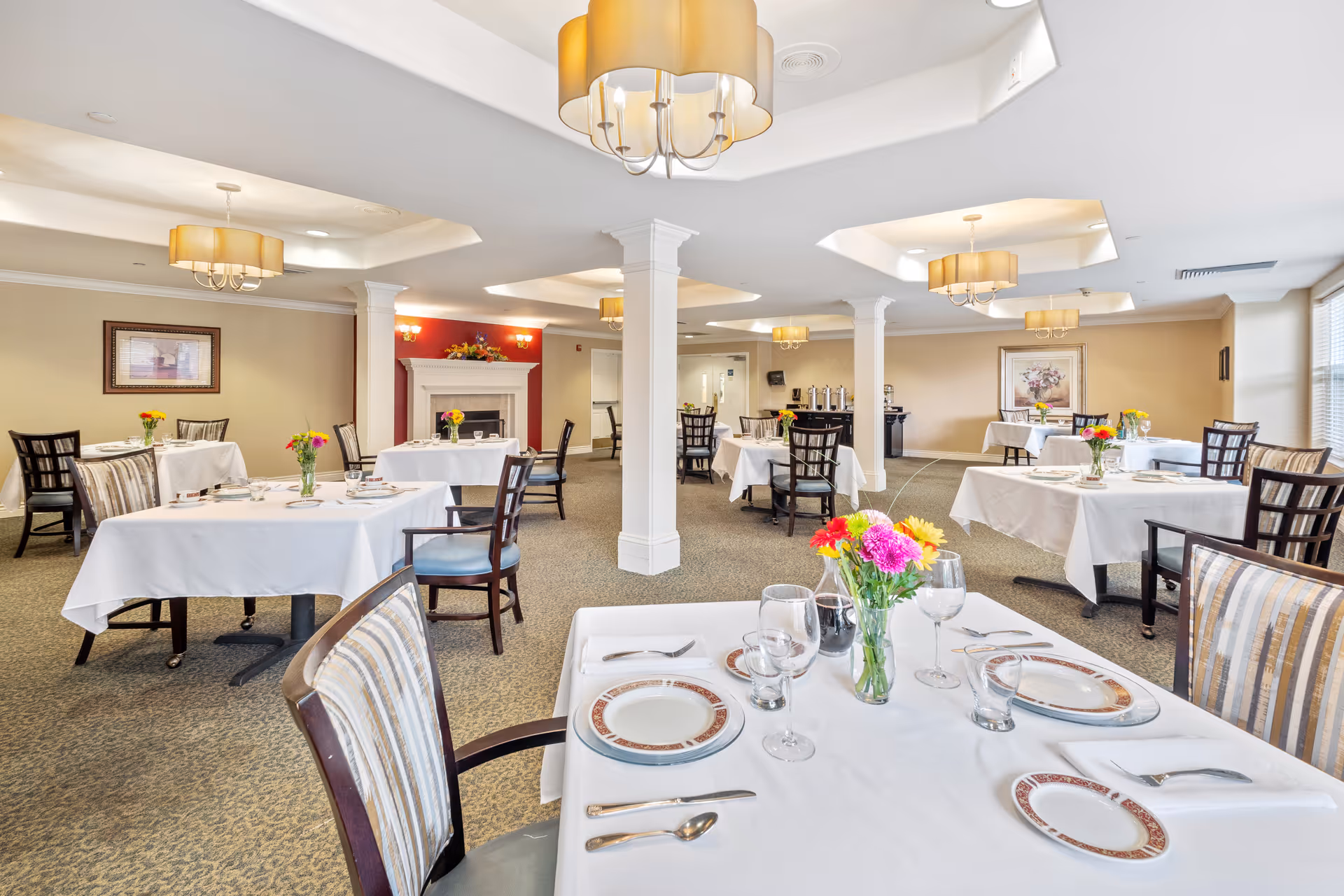 A bright and elegant dining room with multiple tables covered in white tablecloths, each set with plates, glasses, and silverware. Fresh flower arrangements are placed on each table. The room features beige walls, carpeted floors, decorative ceiling lights, and a fireplace with a floral arrangement above it.