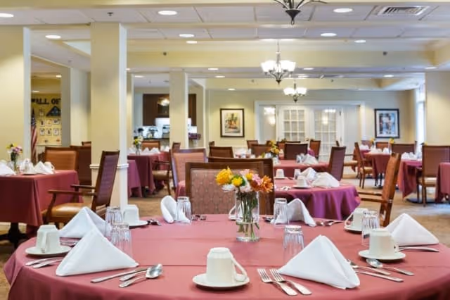 A spacious dining room with multiple round tables covered in maroon tablecloths, each set with white napkins, cups, glasses, and silverware. There are floral centerpieces on the tables, and the room features soft yellow walls, chandeliers, and framed artwork.