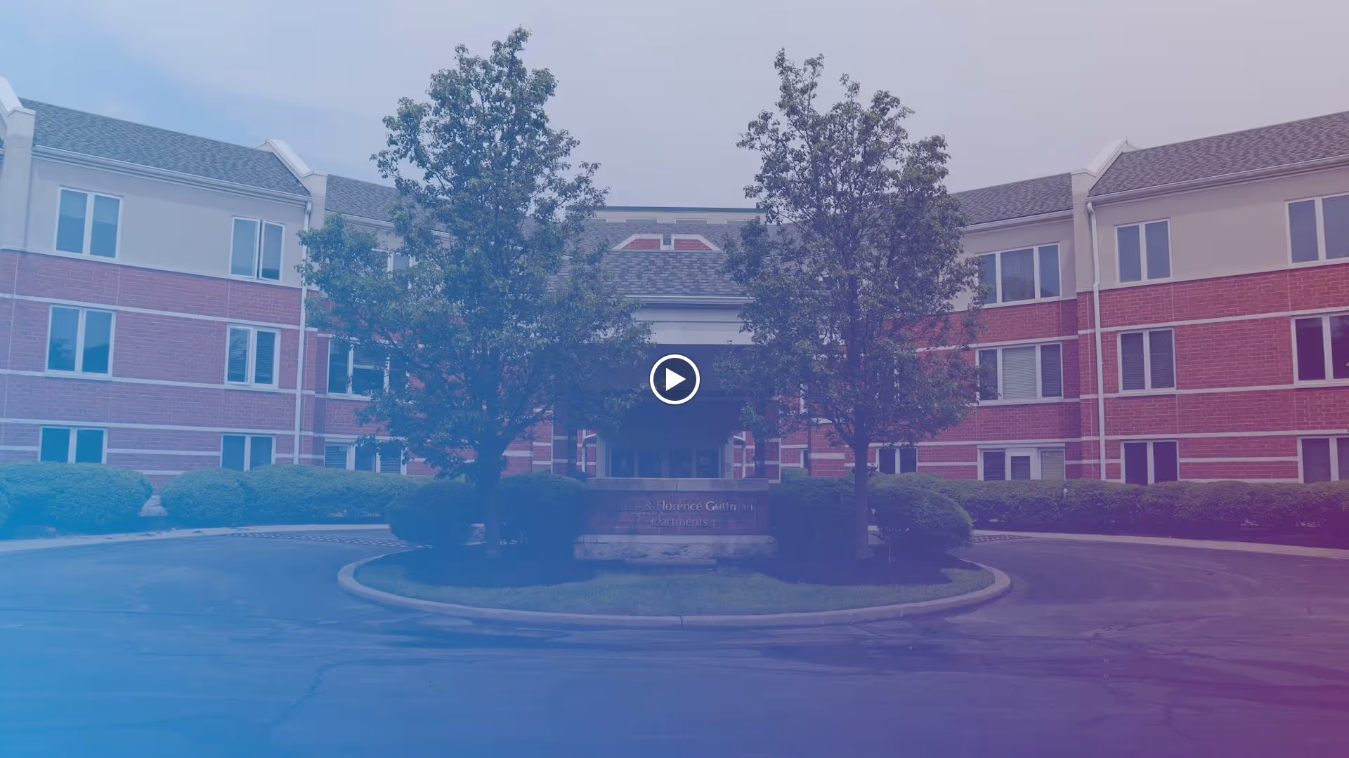 Front exterior view of a multi-story senior living facility with red brick and white trim, featuring a circular driveway with two trees and bushes in the center.