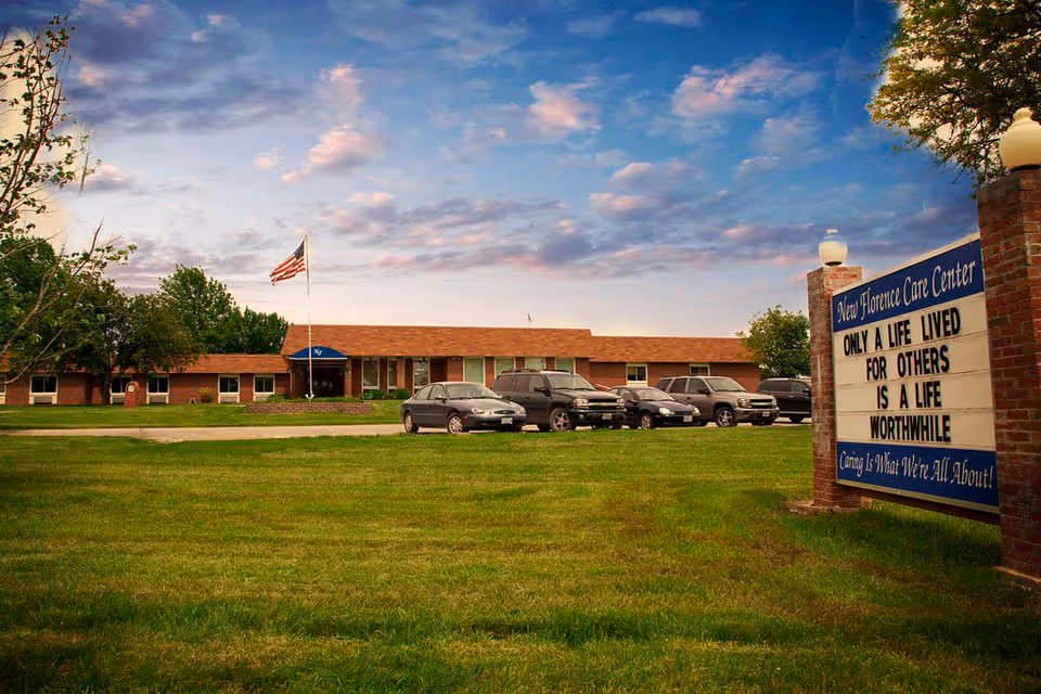 Exterior view of New Florence Care Center, a single-story brick building with a brown roof, several parked cars in front, an American flag on a flagpole, and a sign that reads 'ONLY A LIFE LIVED FOR OTHERS IS A LIFE WORTHWHILE Caring Is What We're All About!' under a partly cloudy sky.