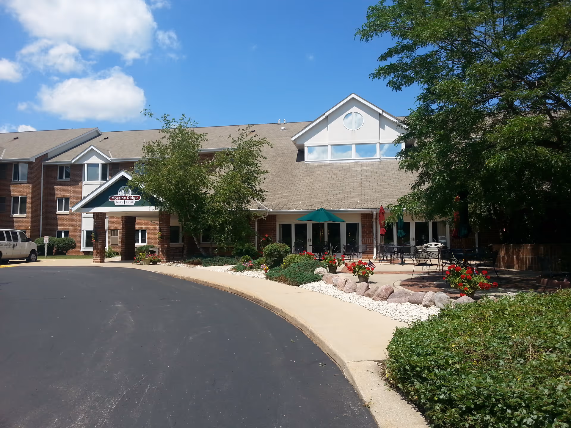 Exterior view of Moraine Ridge Senior Living building on a sunny day with a clear blue sky. The building has a brick facade with multiple windows and a covered entrance. There is a paved driveway and a landscaped area with bushes, flowers, and trees. Outdoor seating with tables and umbrellas is visible on the right side.
