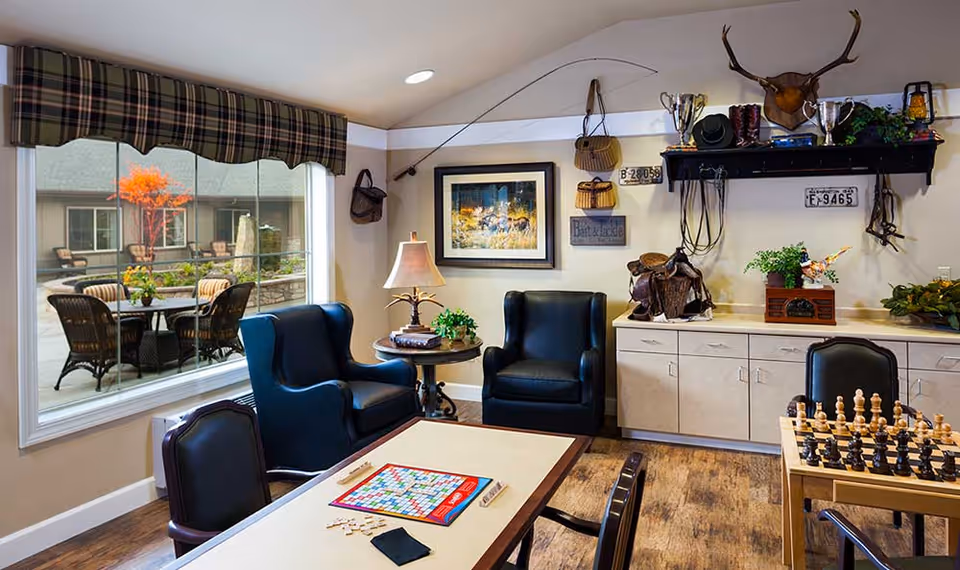 A cozy common area with two black armchairs and a table with a Scrabble game set up. There is a large window showing an outdoor patio with chairs and a small tree with orange leaves. The walls are decorated with western-themed items including a mounted deer head, trophies, baskets, and framed artwork. A chessboard is also visible on a side table.