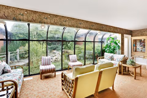 A bright and cozy senior living facility common area with large curved glass windows offering a view of a lush garden outside. The room features floral and striped upholstered chairs and sofas arranged around a small wooden table, with a large green plant in the corner and warm beige carpeting.
