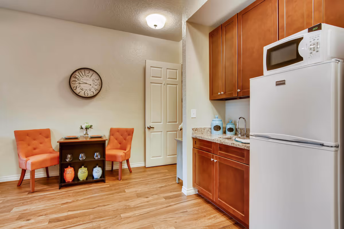 A small kitchen and dining area featuring wooden cabinets, a white refrigerator with a microwave on top, a granite countertop with a sink, and two orange upholstered chairs around a small wooden table. The table has a small flower vase and two plates. A round wall clock is mounted on the beige wall above the chairs, and the floor is wood.