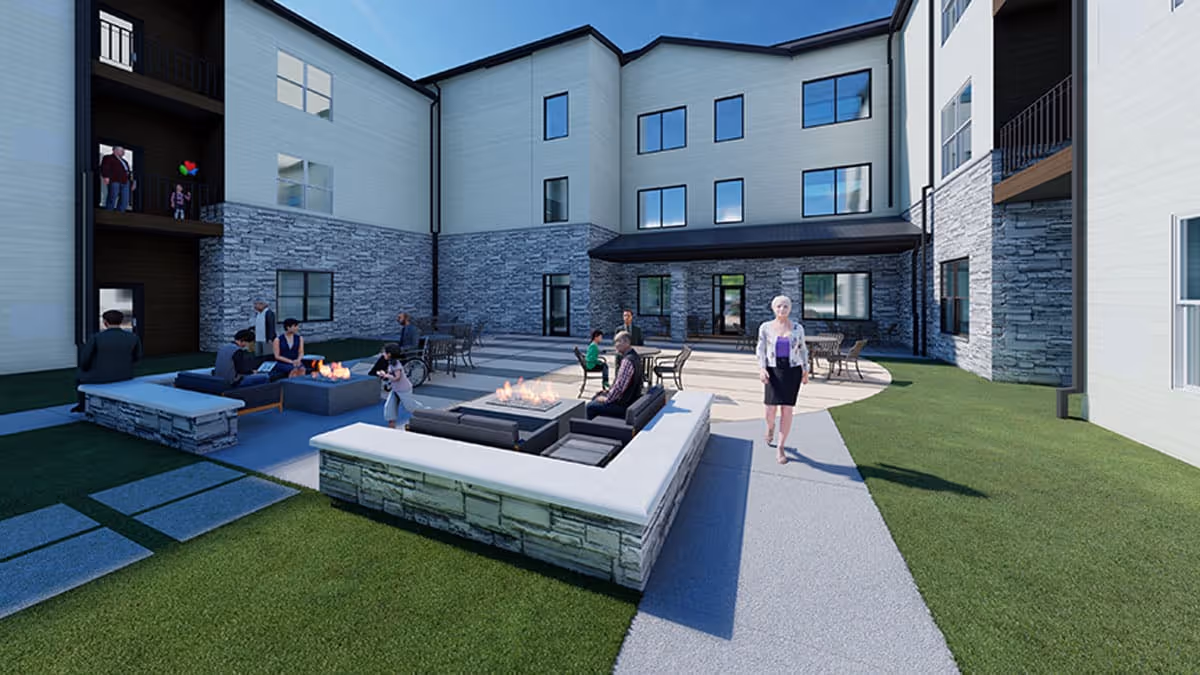 Outdoor courtyard area of a senior living facility with several people sitting and standing around modern stone fire pits and patio furniture. The building surrounding the courtyard has multiple windows and a stone facade on the lower level. The sky is clear and blue.