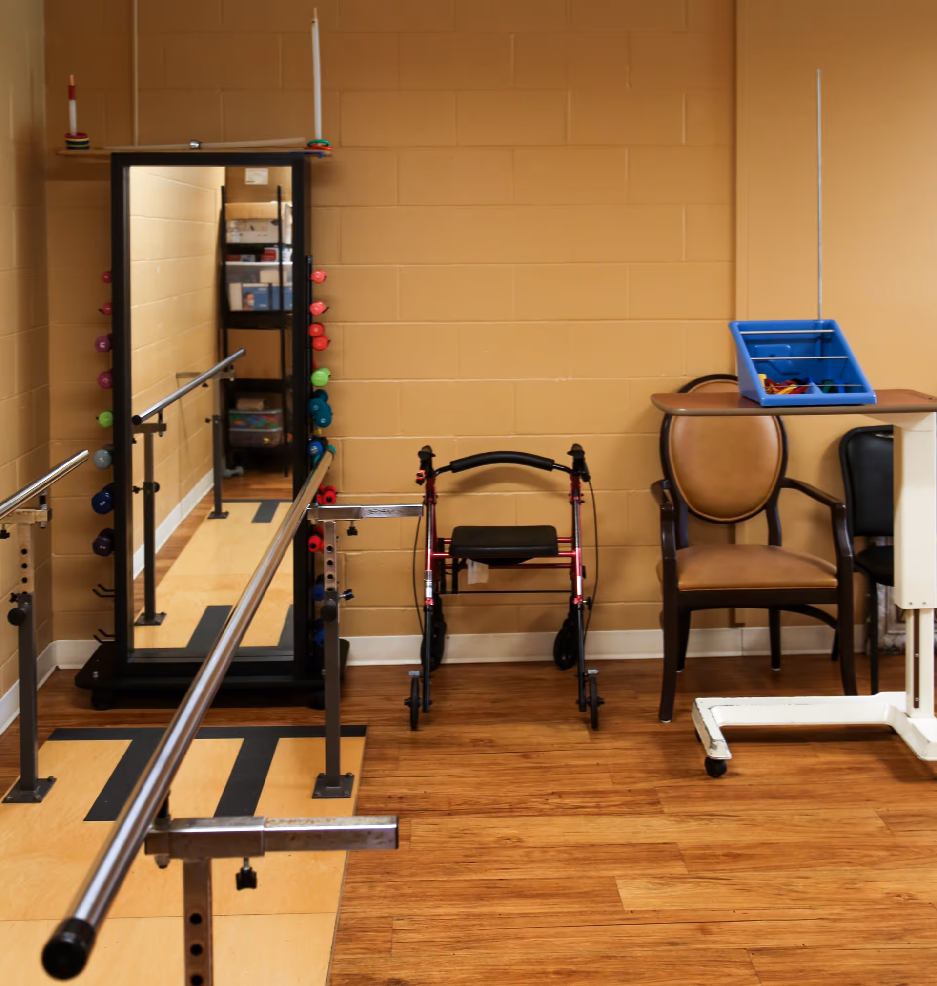 A therapy/exercise room with parallel walking bars, a full-length mirror, a walker, chairs, and small weights against a tan cinderblock wall.