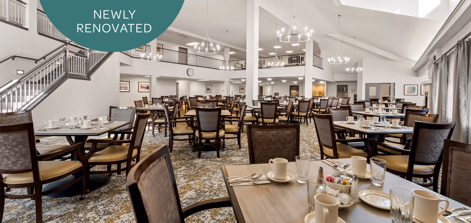 A spacious, newly renovated dining room with multiple wooden tables and chairs arranged neatly. Each table is set with cups, glasses, plates, and silverware. The room features high ceilings, modern chandeliers, large windows with curtains, and a staircase leading to an upper level.
