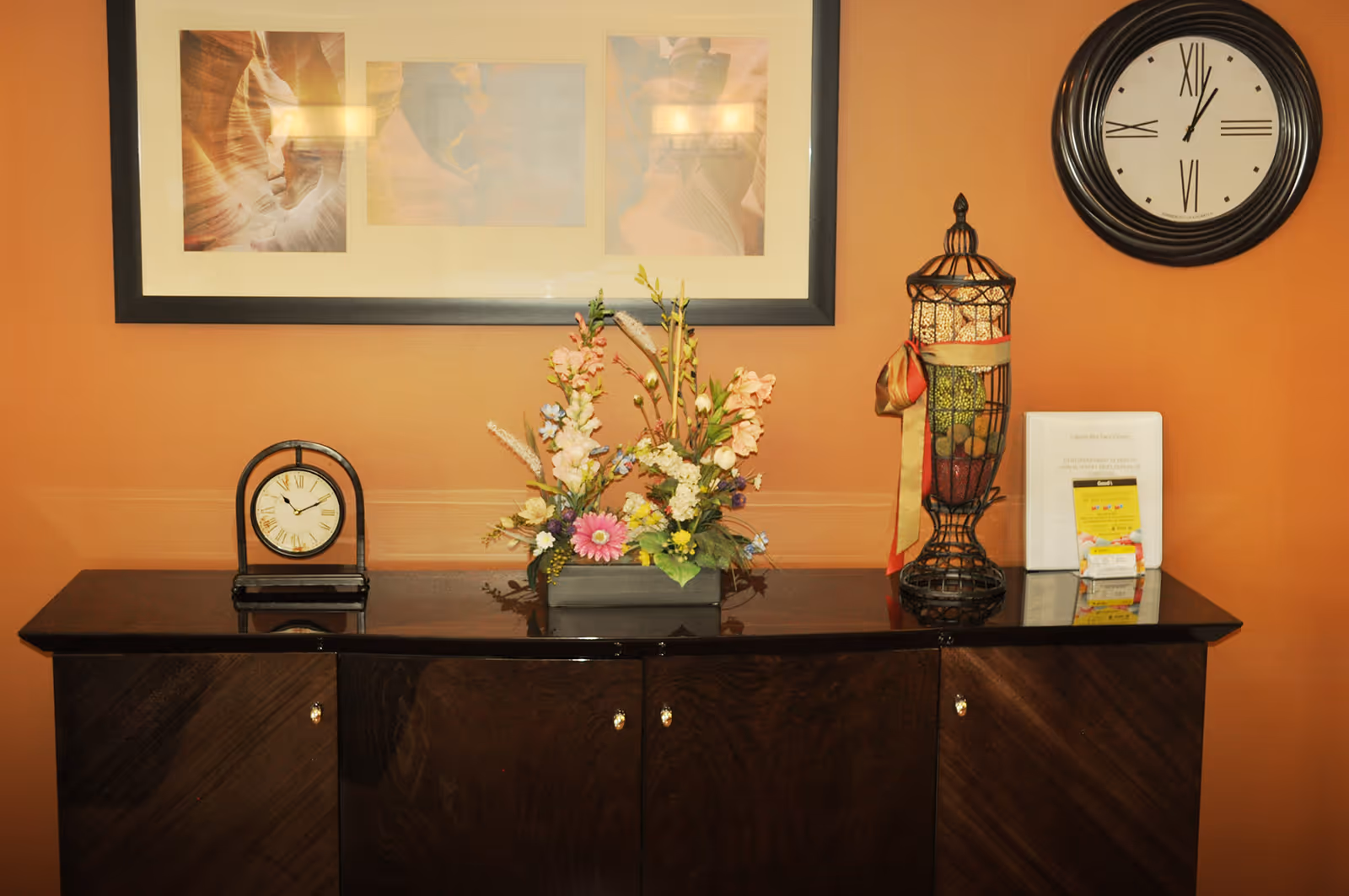 A dark wooden cabinet against an orange wall with a framed triptych photo above it. On the cabinet are a small clock, a floral arrangement in a rectangular vase, and a decorative wire container filled with colorful items and tied with a ribbon. A large round wall clock is mounted on the wall to the right.