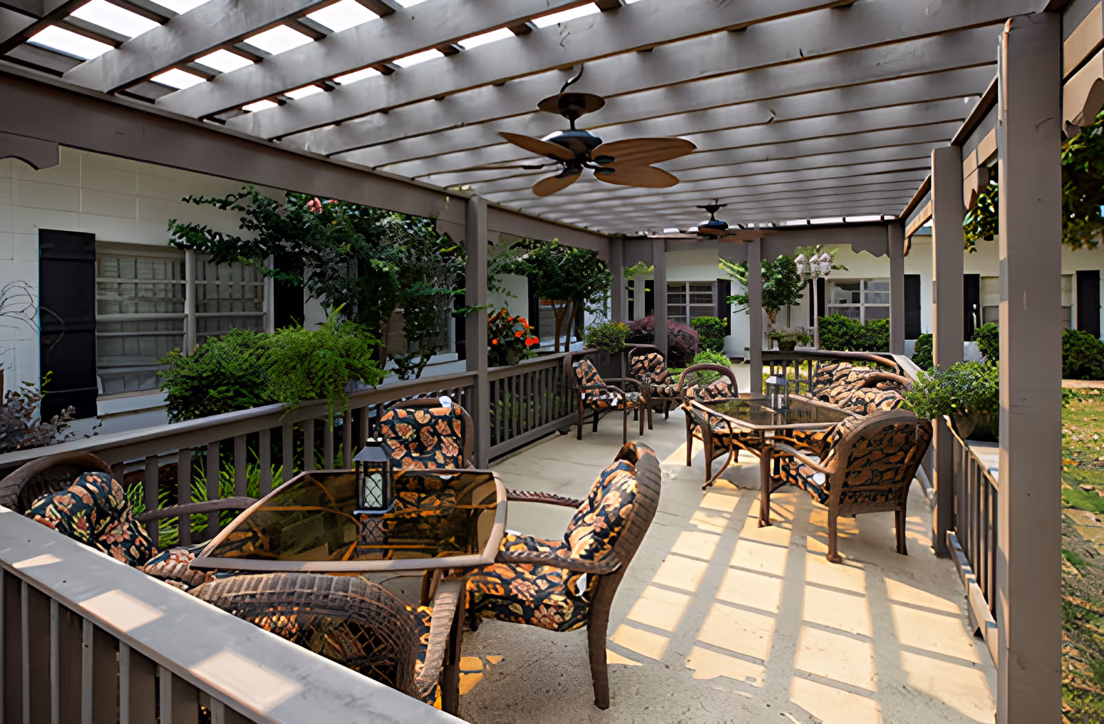 Outdoor covered patio area with cushioned wicker chairs and glass-top tables, ceiling fans, and surrounding greenery at Ahc West Tennessee Transitional Care.