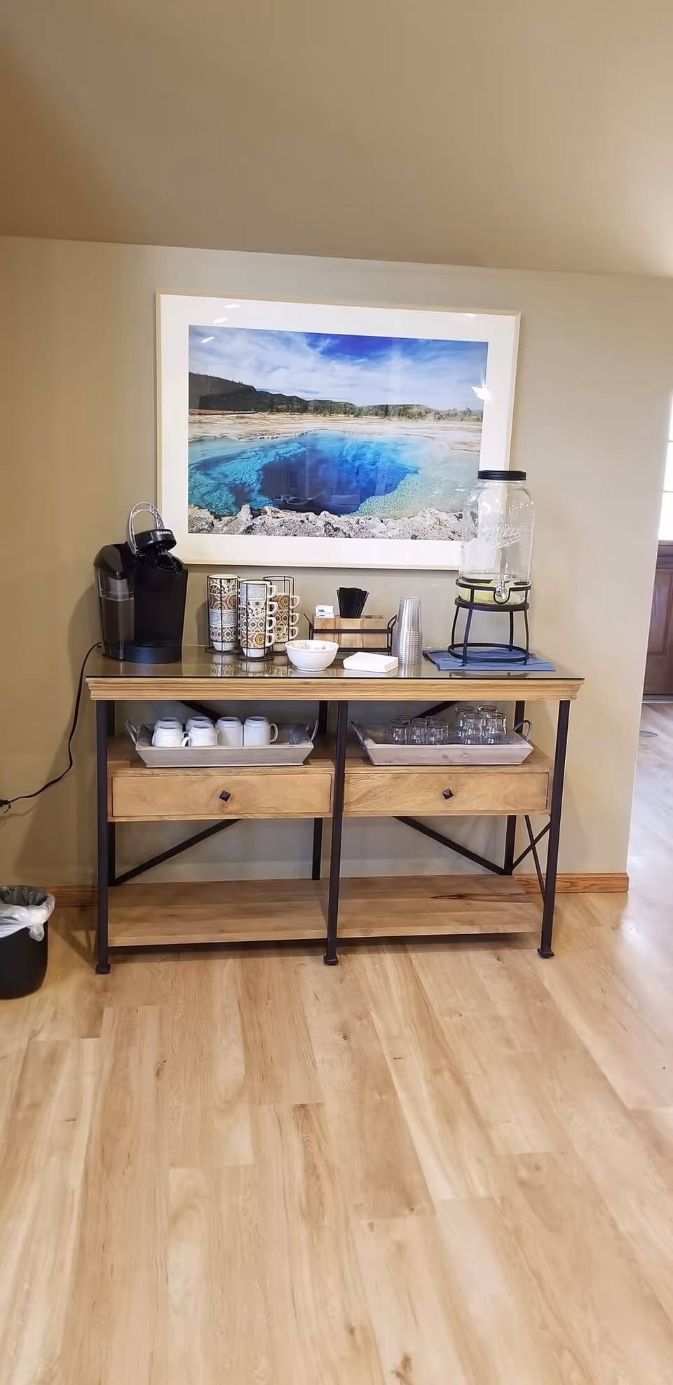 A wooden console table with a coffee maker, mugs, glasses, a water dispenser, and napkins arranged on top. Above the table is a framed photograph of a natural hot spring with clear blue water and a scenic background. The floor is light wood, and the walls are beige.