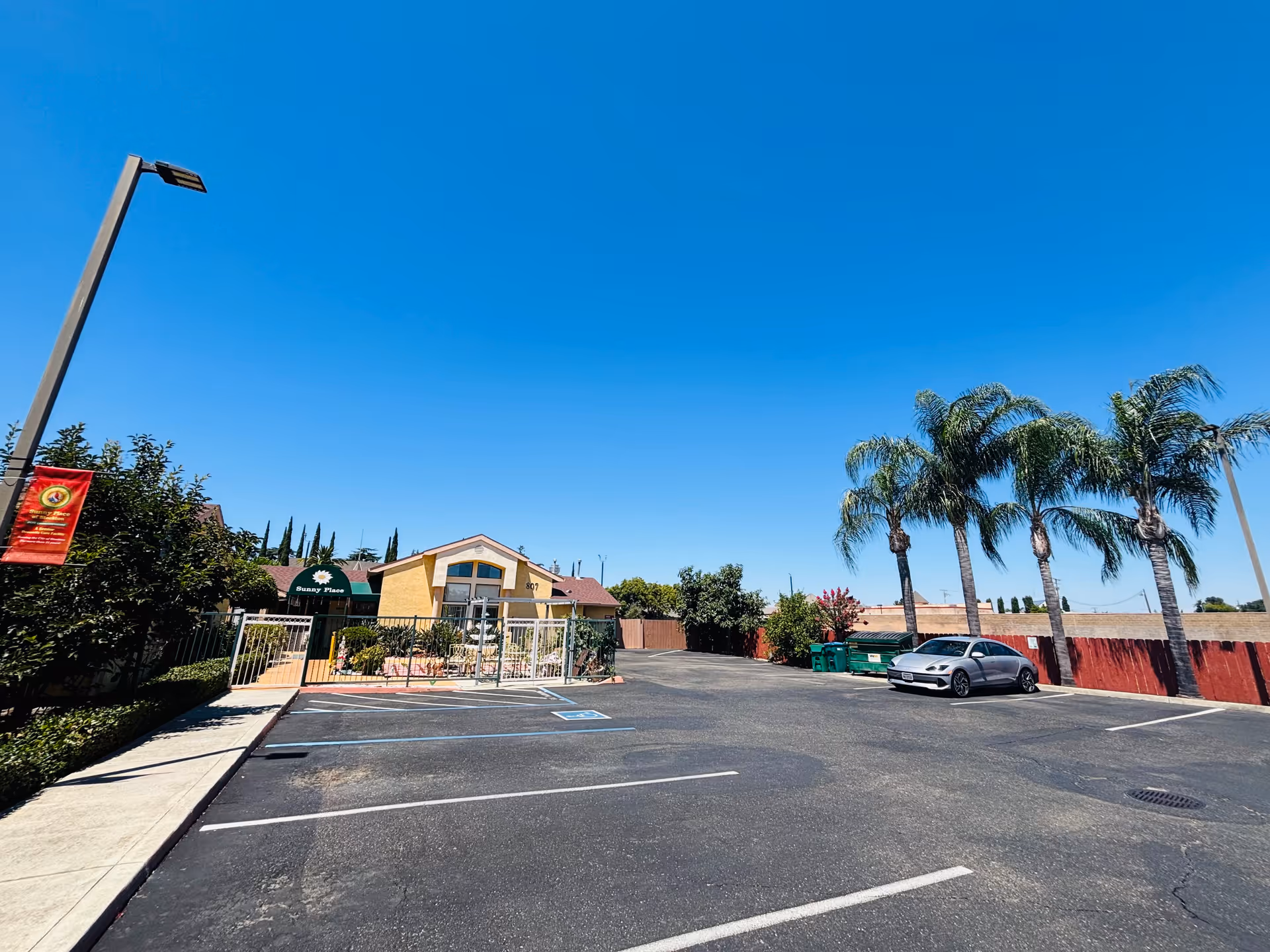 Parking lot and entrance of Sunny Place of Stockton with a small yellow building, palm trees, and a clear blue sky.